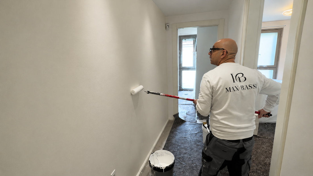 Max Bassi dipinge con il rullo a manico lungo la parete di un corridoio, con secchio di pittura bianca e telo di protezione a terra